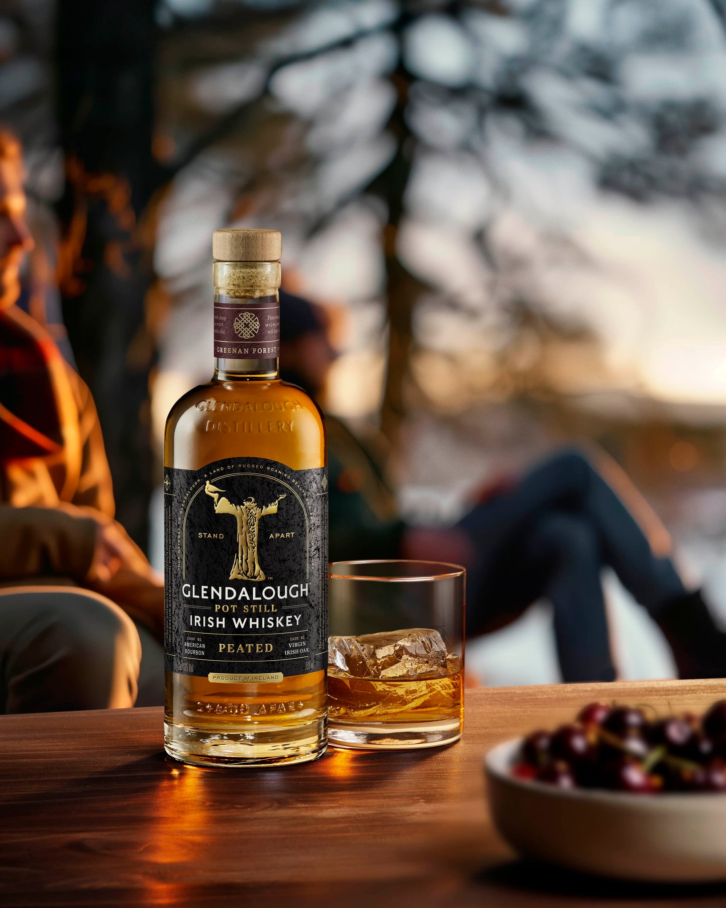 A bottle of Glendalough Peated Irish Whiskey and a glass with ice on a wooden table, with a blurred outdoor background.
