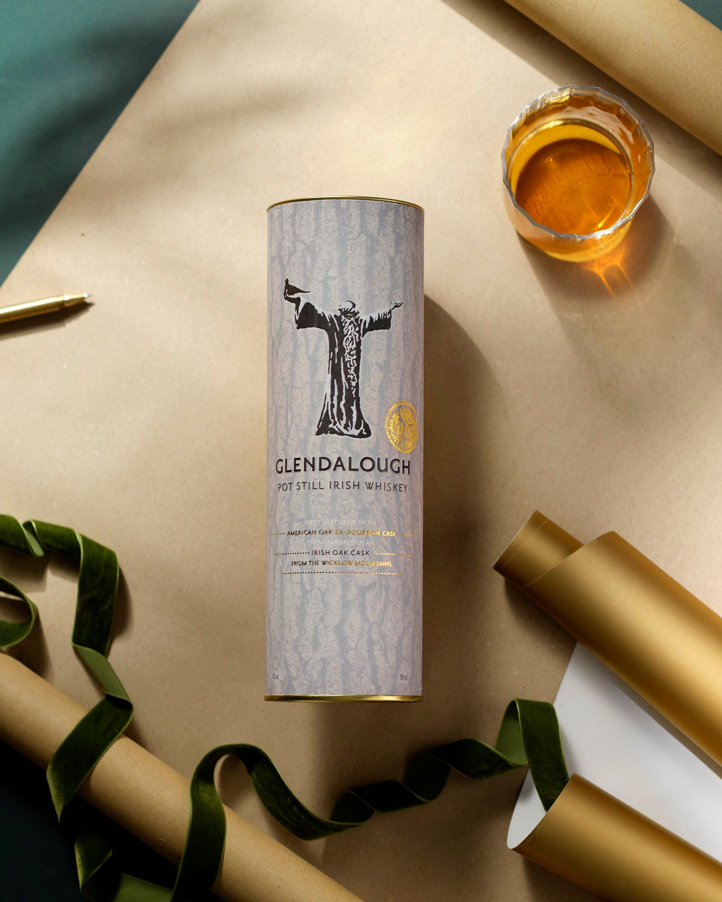 A Glendalough Pot Still Irish Whiskey gift tube surrounded by gold wrapping paper rolls, a green velvet ribbon, and a glass of whiskey.