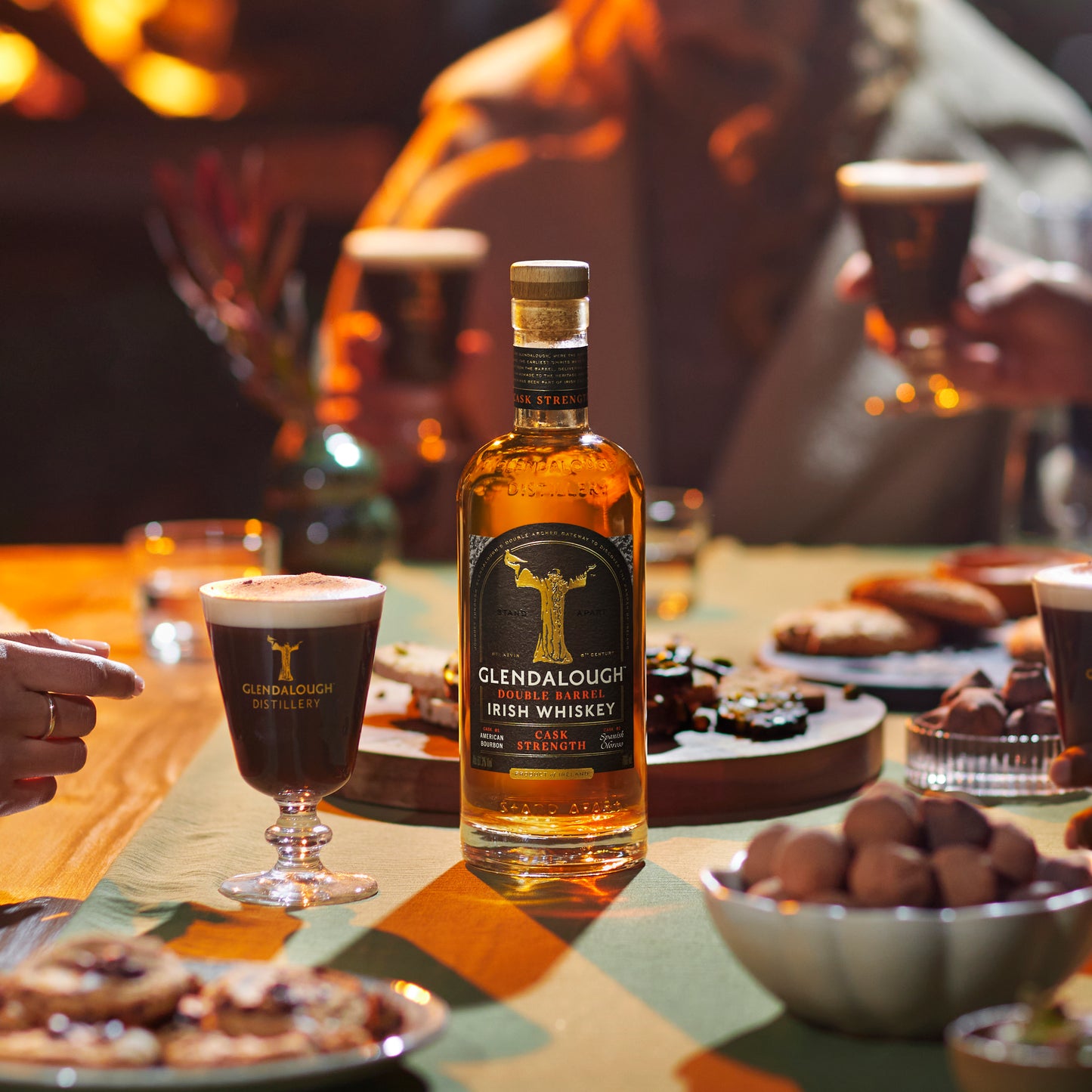A bottle of Glendalough Cask Strength Irish Whiskey sits on a table surrounded by Irish coffees, snacks, and people